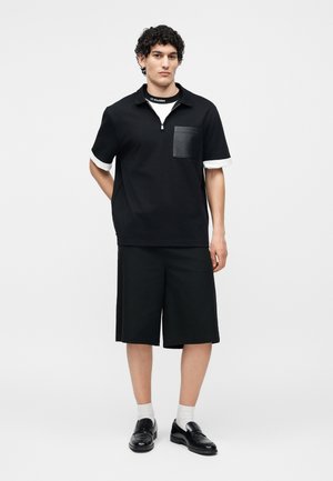 Nuori mies kiharatukkaisena, pukeutuneena mustaan lyhythihaiseen paitaan, jossa on etutasku, mustiin polvipituisiin shortseihin, valkoisiin sukkiin ja mustiin kenkiin.