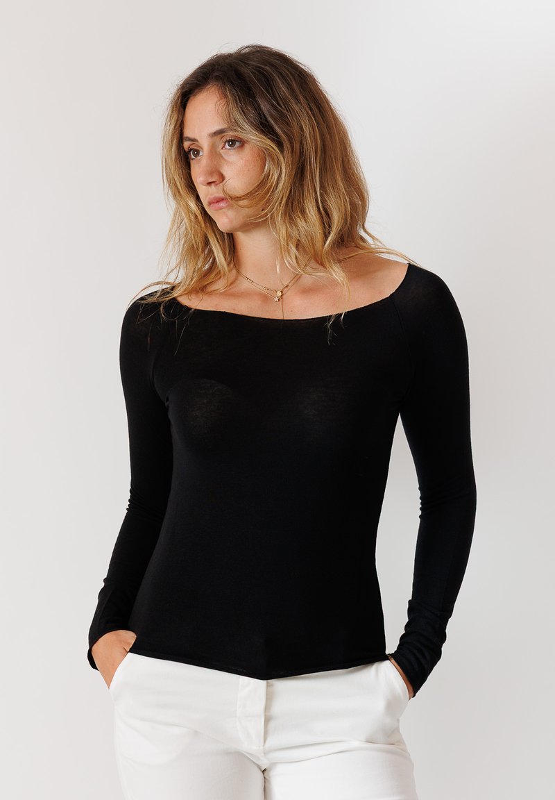 Maglietta nera a maniche lunghe con scollatura quadrata, design aderente e tessuto morbido, abbinata a pantaloni bianchi. Il modello è in piedi con le mani in tasca.