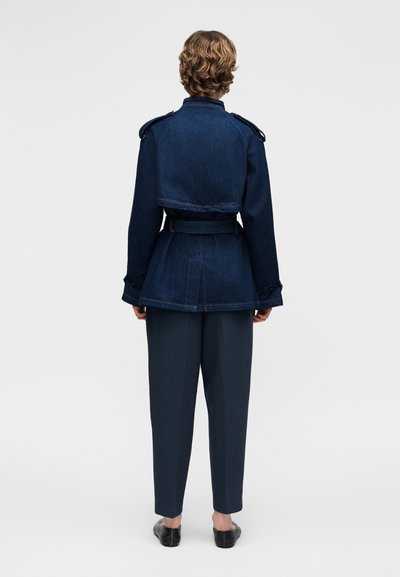 Person med kort krøllet hår står med ryggen mod, iført en mørkeblå jakke med bælte og epauletter samt mørke, skræddersyede bukser og sorte sko.