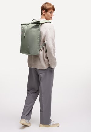 Joven con suéter beige y pantalones grises lleva una mochila grande de color verde claro, de pie frente a un fondo blanco liso.