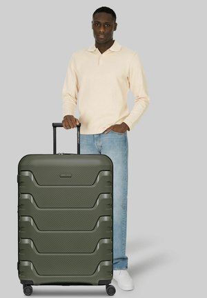 Homme en chemise beige à manches longues et jean, debout avec la main sur la poignée d'une grande valise rigide verte sur fond gris.