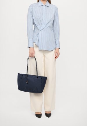 Femme portant un chemisier à rayures bleues et un pantalon crème, tenant un grand sac cabas bleu marine avec des poignées en chaîne et des chaussures noires pointues.