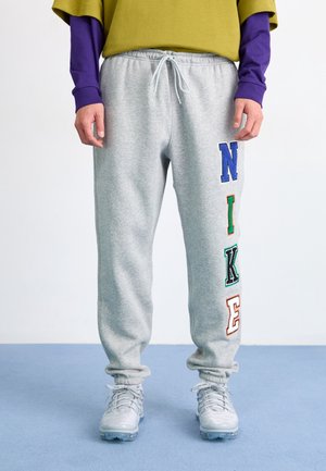 Osoba ubrana w szare spodnie dresowe z dużymi, wielokolorowymi literami "NIKE" na prawej nodze, białe trampki, fioletową bluzę z długim rękawem oraz oliwkowo zielony top.