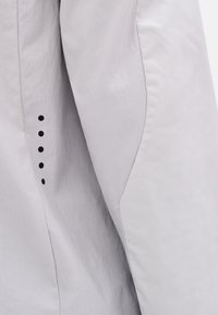 Hellgrauer Jackenärmel mit glatter Textur, Nahtdetails und sechs schwarzen Zierknöpfen entlang der Kante.