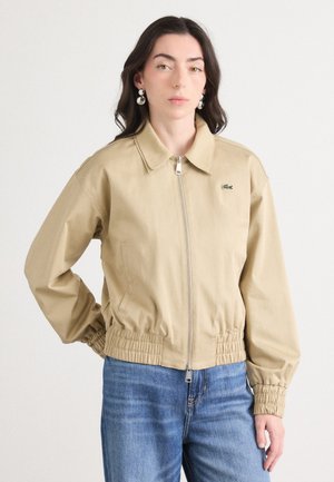 Beige jakke med krage, glidelås foran og elastisk midje. Har lange ermer og en liten grønn logo på venstre bryst.