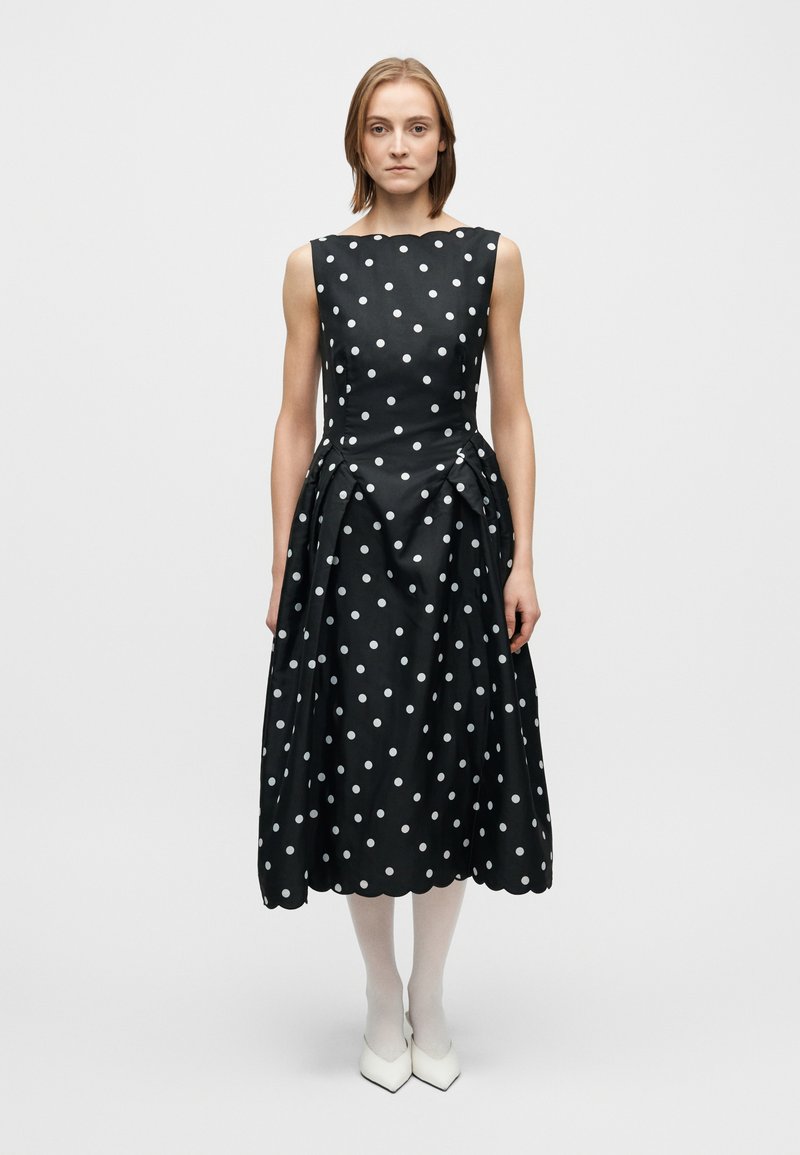 Svart klänning med vita prickar, tajt överdel, utsvängd kjol och sidoskicklade veck. Tillverkad av glansigt tyg. Modellen bär vita strumpbyxor och spetsiga klackskor.