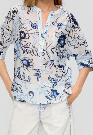 Vrouw draagt een witte blouse met blauwe bloemmotieven en een voorzak, gecombineerd met witte broek, handen in de zakken.