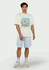 Valkoinen puuvillapaita, jossa on värikäs kaupunkimaisema-prints, yhdistettynä vaaleansinisiin shortseihin. Sneakerit ja sukat täydentävät rentoa ilmettä.