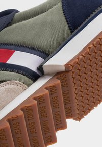 Sneakers in tessuto verde con dettagli blu navy, una striscia rossa e bianca, e una suola in gomma testurizzata color cuoio con logo in rilievo.