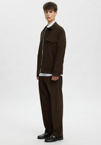 Brun knappet jakke med brystlomme, kombinert med løstsittende brune bukser og blanke svarte loafers over en lys stripet skjorte.