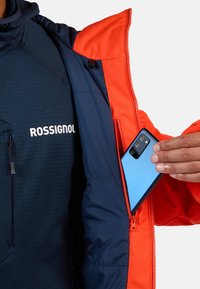 Blauw en oranje jack met ritszakje voor een smartphone. Tekst "ROSSIGNOL" op marineblauwe stof; gladde, gevoerde textuur, functioneel ontwerp.
