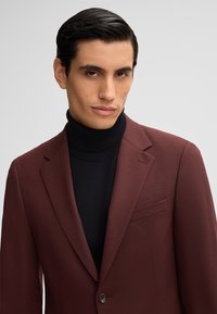 Blazer tailored color mogano con rever a punta e una sottile texture, indossato sopra un maglione nero a collo alto. Presenta una tasca frontale e chiusura con bottone.