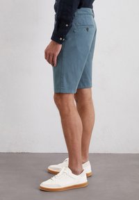 Heren shorts in blauwe katoen, knielang, met zijzakken en een achterzak met knoop. Gecombineerd met witte sneakers, gezien vanaf de zijkant.