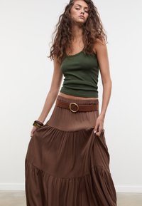 Jupe longue marron à volants avec une silhouette évasée, associée à un débardeur vert ajusté. Une large ceinture marron avec une boucle dorée souligne la taille.