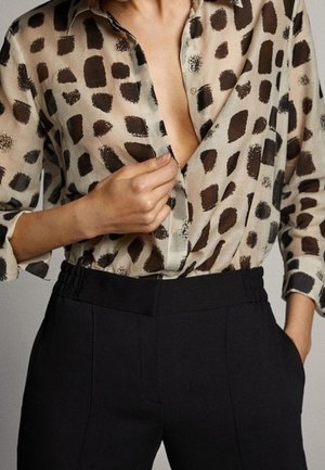 Person trägt ein beiges Hemd mit braunen quadratischen Mustern, knöpft das Hemd zu, dazu trägt die Person schwarze, hoch taillierte Hosen mit elastischem Bund.