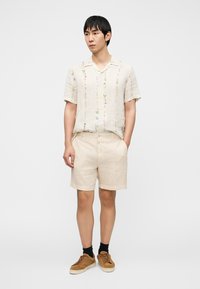 Ελαφρύ πουκάμισο με κοντό μανίκι από λινό με κεντήματα με λουλούδια σε παστέλ χρώματα, συνδυασμένο με μπεζ λινό σόρτς και καφέ αθλητικά παπούτσια.