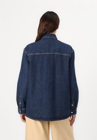 Jachetă din denim de culoare albastru închis, cu o guler ascuțit, mâneci lungi și manșete cu nasturi. Textură netedă cu cusături vizibile și o croială lejeră.