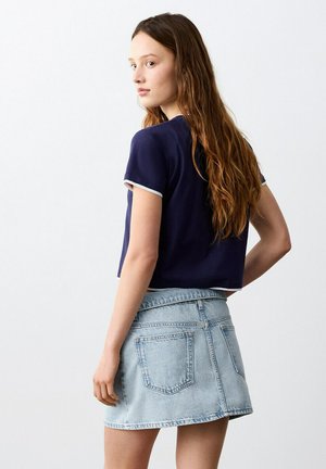 Giovane donna con lunghi capelli castani che indossa una maglietta blu navy a maniche corte e una minigonna di denim chiaro, leggermente girata di lato.