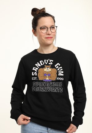 Kvinde med briller iført en sort sweatshirt, der viser muskuløse Sandy Cheeks fra SpongeBob SquarePants og teksten "Sandy's Gym Est. 1999".