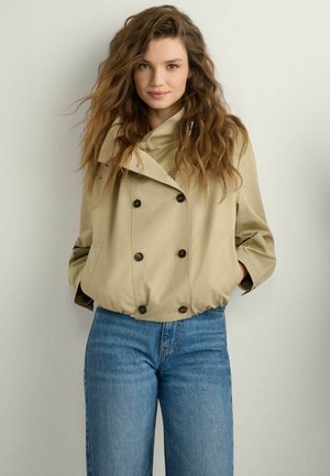 Kobieta z długimi falującymi włosami, ubrana w beżową dwurzędową kurtkę i niebieskie dżinsy, stojąca na tle gładkiego, jasnego tła.