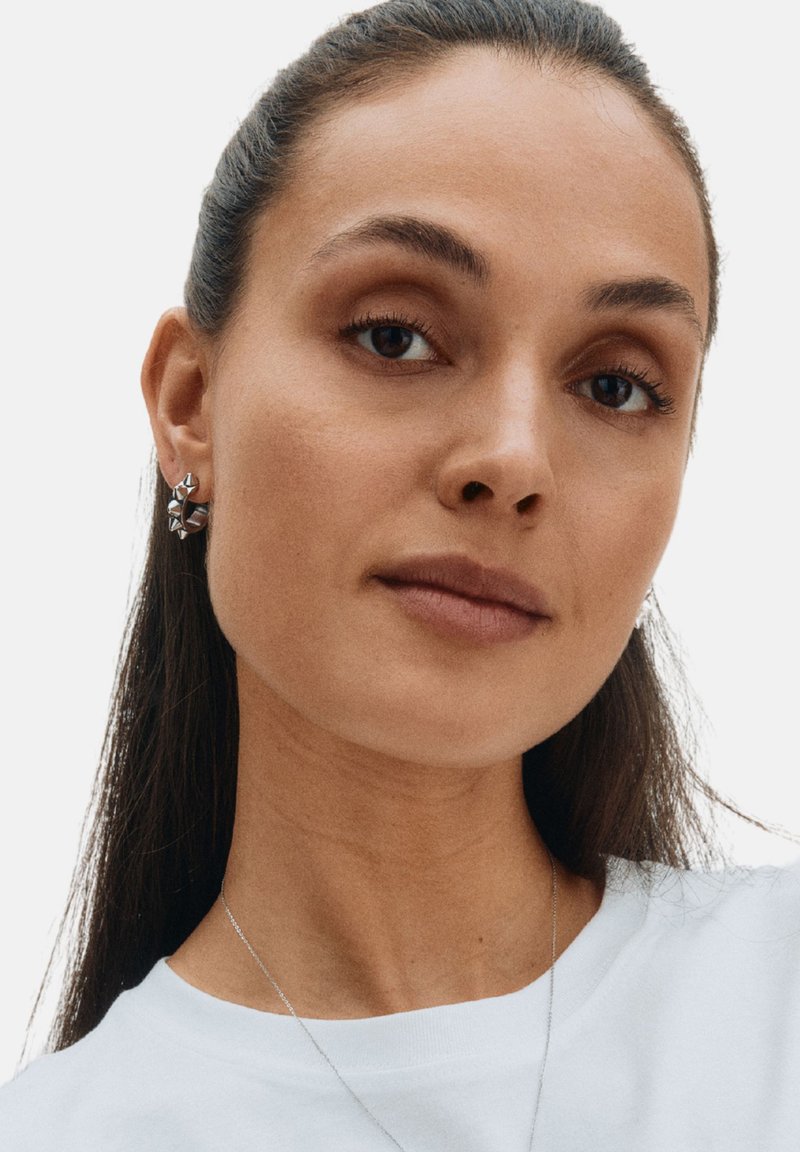 Hõbekõrvarõngad geomeetrilise kujundusega, kus on nurgelised kujundid ja poleeritud viimistlus, kantud modellil, kellel on pikad juuksed ja valge särk.