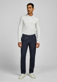 Man in een witte lange mouw polo, donkere blauwe broek en witte sneakers, staand tegen een effen grijze achtergrond.