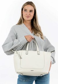 Sac à main blanc texturé avec des accents noirs et des poignées robustes, présentant un design spacieux et une marque discrète. Le modèle porte une chemise rayée.