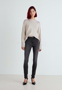 Jeans grises ajustados combinados con un suéter de punto beige claro, que presenta puños acanalados, y tacones negros brillantes, sobre un fondo neutro.