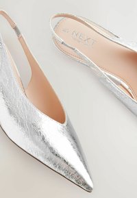 Zapatos de tacón alto plateados, tipo slingback, con punta afilada, presentando un acabado metálico texturizado y una plantilla nude con la marca "NEXT".