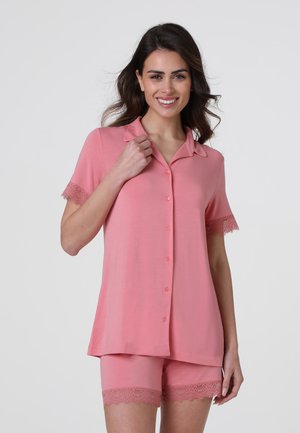 Donna sorridente, indossa una maglietta rosa a maniche corte con bottoni e pantaloncini abbinati con rifiniture in pizzo sulle maniche e sull'orlo su sfondo uniforme.