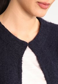 Tmavě modrý texturovaný cardigan s kulatým výstřihem a bez zapínání, vrstvený přes světle zbarvený top. Tkaniny vypadají měkké a huňaté.