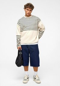 Creme sweatshirt met zwarte horizontale strepen, gecombineerd met losse blauwe denim shorts. Accessoires zijn een zwarte tas en witte sokken met sneakers.