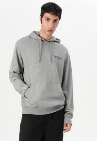Felpa grigia con cappuccio, tasca frontale, coulisse e dettaglio logo nero sul petto. Tessuto morbido con una vestibilità comoda.