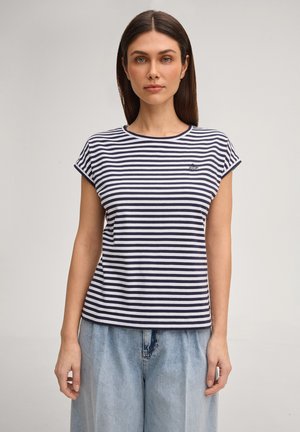 Kobieta ubrana w granatowo-biały prążkowany krótki rękaw i jasnoniebieskie spodnie z wysokim stanem, stojąca na jednolitym tle.