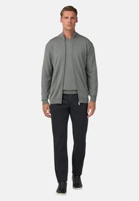 Světlé šedé zipové mikina s vysokým límcem, v kombinaci s odpovídajícím šedým tričkem s kulatým výstřihem a černými kalhotami, doplněné černými teniskami.