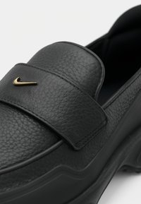 Μαύρο δερμάτινο παπούτσι slip-on με υφή, παχύ πάτο και χρυσό λογότυπο Nike σούσι στο ευρύ λουρί που διασχίζει το πάνω μέρος.