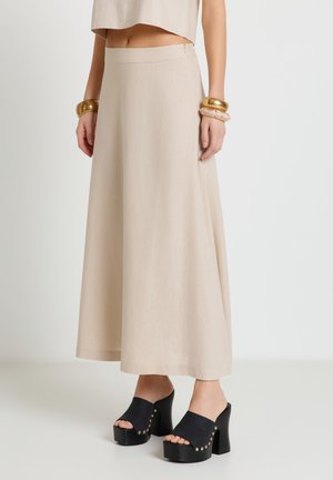 Frau trägt einen beigefarbenen Maxirock mit hoher Taille, schwarze Plateau-Sandalen mit hohen Absätzen und goldenen Nieten sowie klobige goldene Armbänder an beiden Handgelenken.