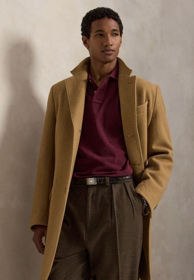 Homme portant un manteau camel sur un polo bordeaux et un pantalon marron à carreaux, debout contre un mur clair uni, une main dans la poche.
