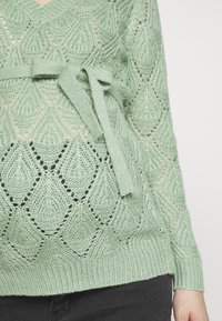 Pull vert clair en tricot avec un motif en losange, doté d'un lien à la taille et de poignets et ourlet côtelés. La texture est aérée et structurée.