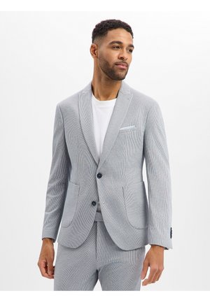 Man draagt een lichtgrijs colbert en bijpassende broek over een wit overhemd, kijkt iets naar zijn rechterkant tegen een effen achtergrond.