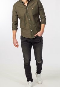 Dark green button-up shirt with a chest logo, paired with slim black jeans and white sneakers. Shirt features long sleeves and a collar.