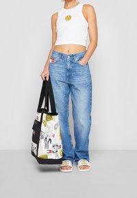 Bílý crop top se smajlíkem, světle modré džíny s vysokým pasem a vzorovaná taška s černými detaily. Bílé pantofle s logem.