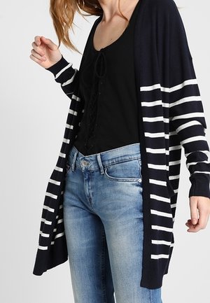 Femme portant un jean bleu, un haut noir à lacets et un long cardigan noir avec des rayures horizontales blanches.