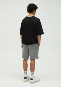 T-shirt noir surdimensionné, shorts en coton gris avec poches, baskets blanches avec accents bleu marine et chaussettes à motifs. Vue de dos.