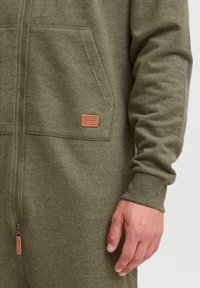 Olive green zip-up onesie in soft fabric, featuring a front pocket and ribbed cuffs. A small leather label is attached near the zipper.