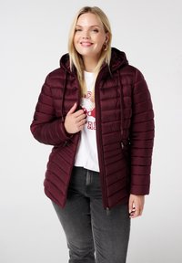 Doudoune bordeaux avec coutures horizontales, fermeture éclair, poches latérales et capuche. Portée par-dessus une chemise blanche et un jean gris.