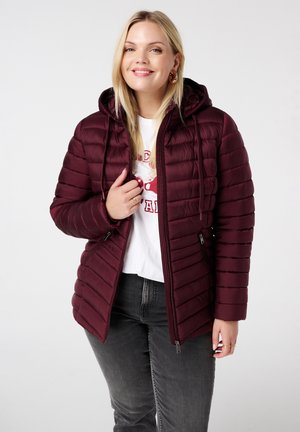 Bordeaux pufferjack met horizontale stiksels, ritssluiting, zijzakken en een capuchon. Geddragen over een wit shirt en grijze jeans.