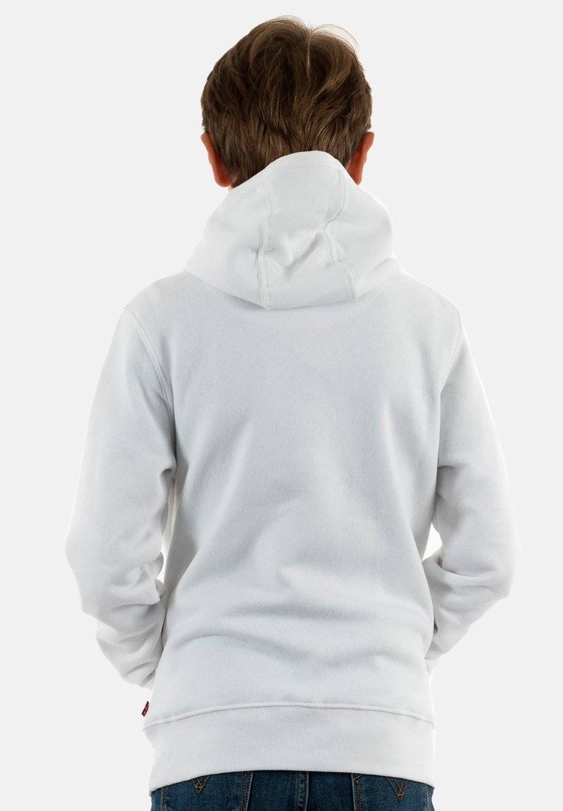Persoon met kort bruin haar, gekleed in een witte hoodie en blauwe jeans, gezien van achteren tegen een effen achtergrond.