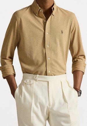 Homme portant une chemise beige boutonnée à manches longues avec un logo brodé et un pantalon plissé crème taille haute, mains dans les poches.