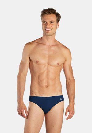 Uomo sorridente in piedi con slip da bagno blu navy, rivolto leggermente a sinistra contro uno sfondo chiaro e semplice, che mostra un fisico atletico e in forma.